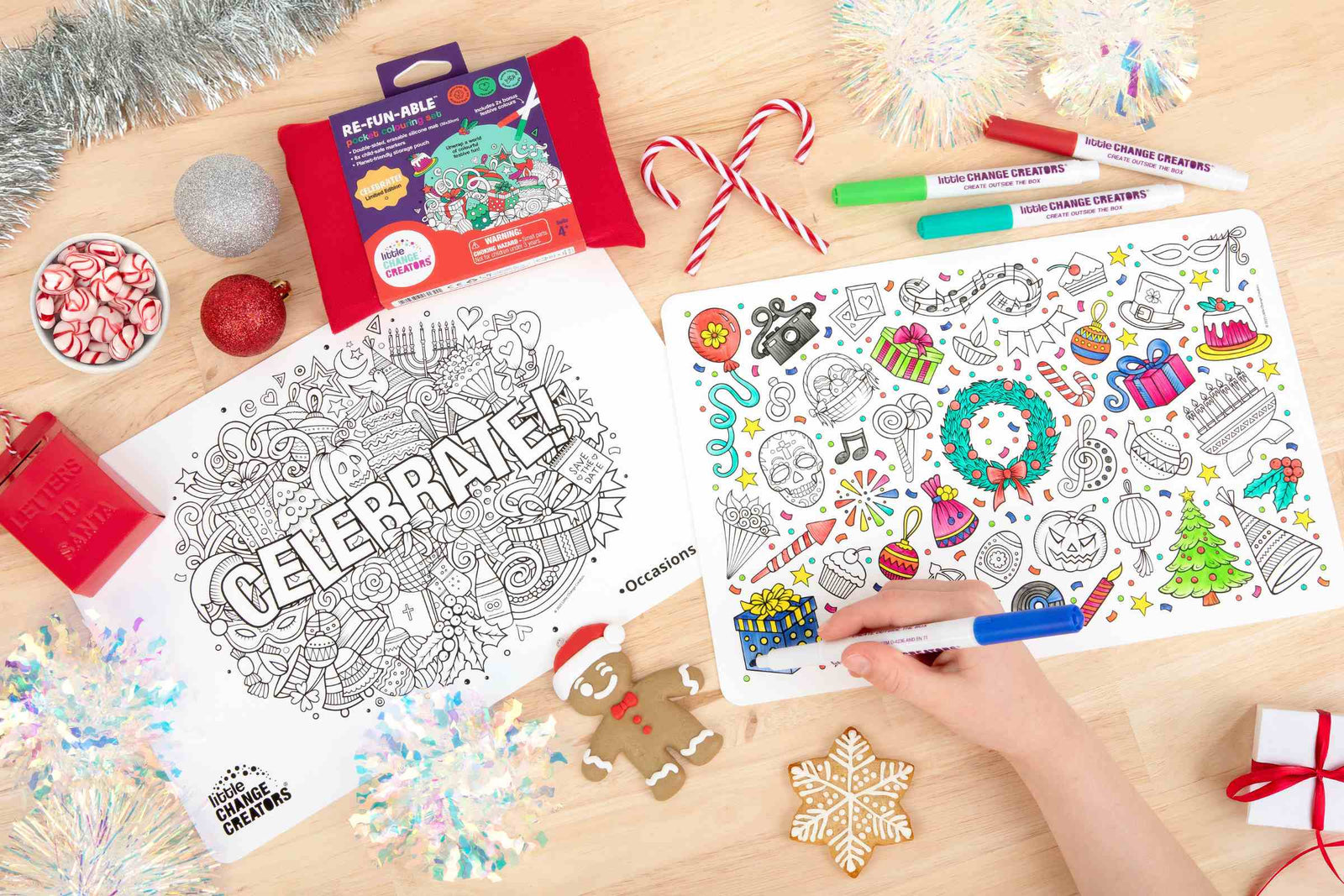 A wooden table top with two silicone colouring mats featuring festive illustrations. The mats are surrounded by Christmas decorations including tinsel and candy canes.