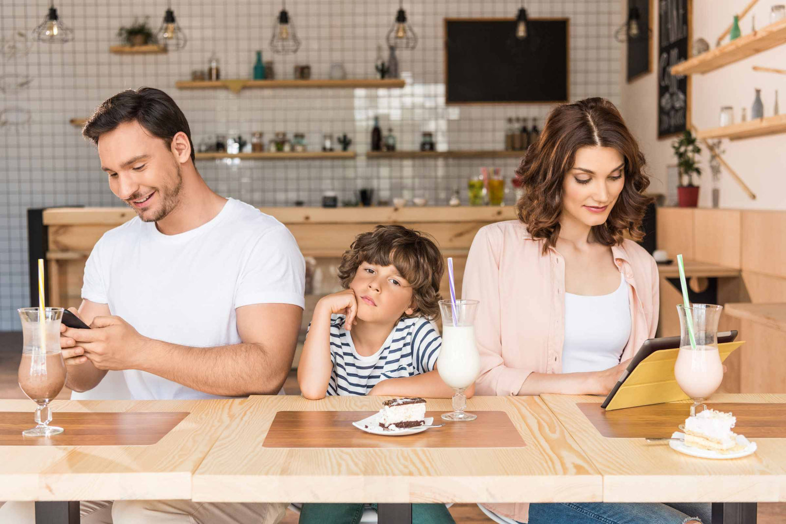 A young boy looking bored as he waits at a restaurant.  His parents are sitting either side of him but they're distracted by their phones.  The image is well-lit with white, beige, peach fuzz tones.