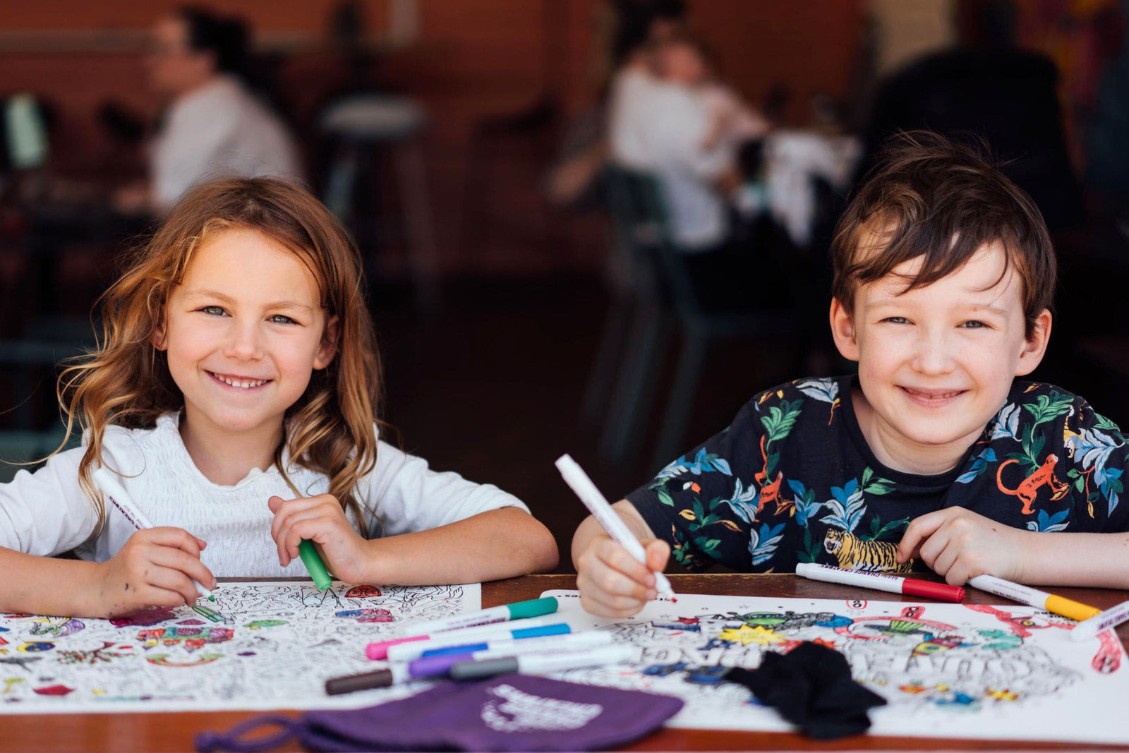 Two children smile as they colour reusable silicone colouring mats at a cafe.