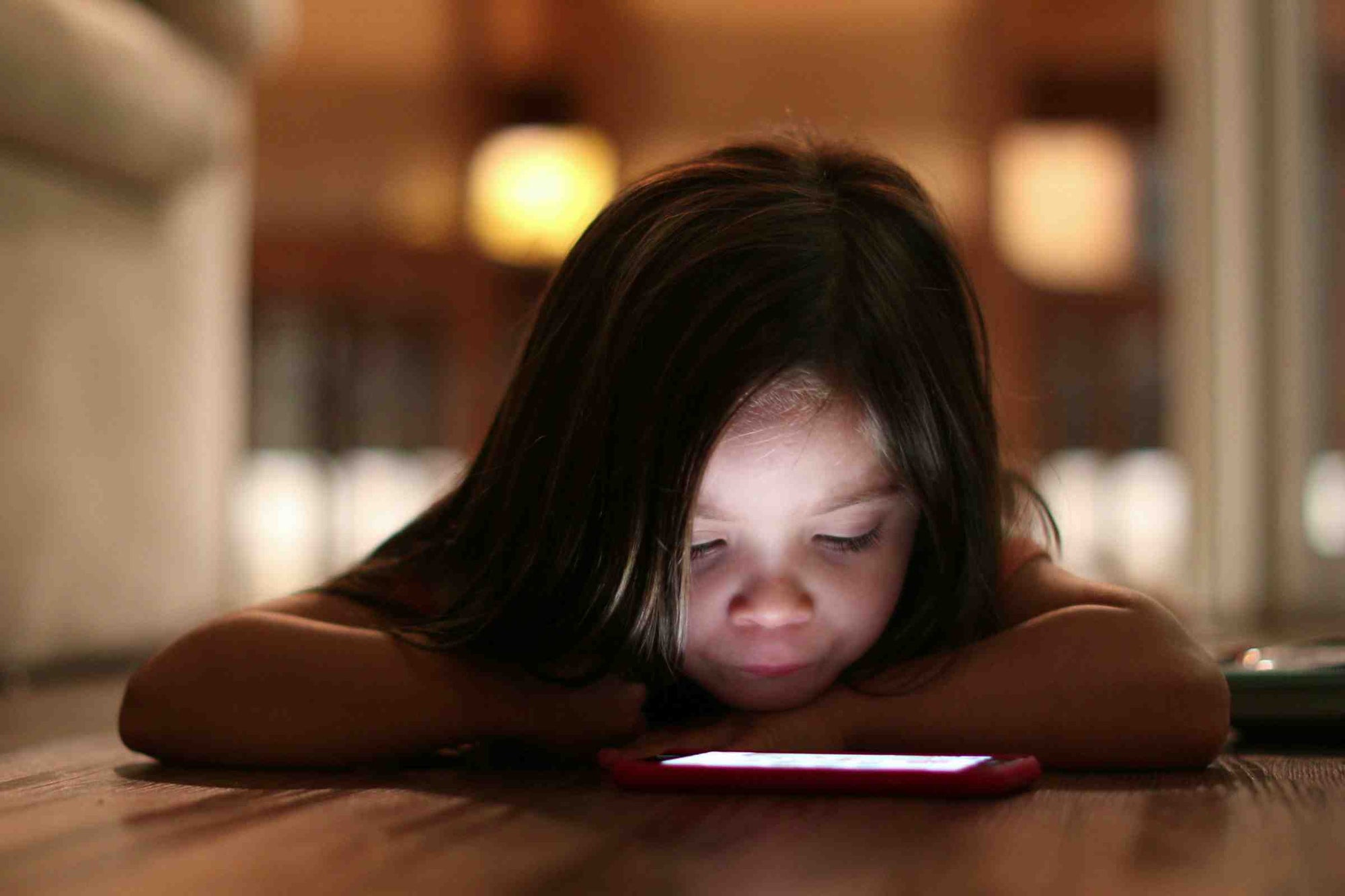 A young girl fixated on a phone with the screen's light illuminating her face.  The image suggests that screentime may impact a child's mental health.