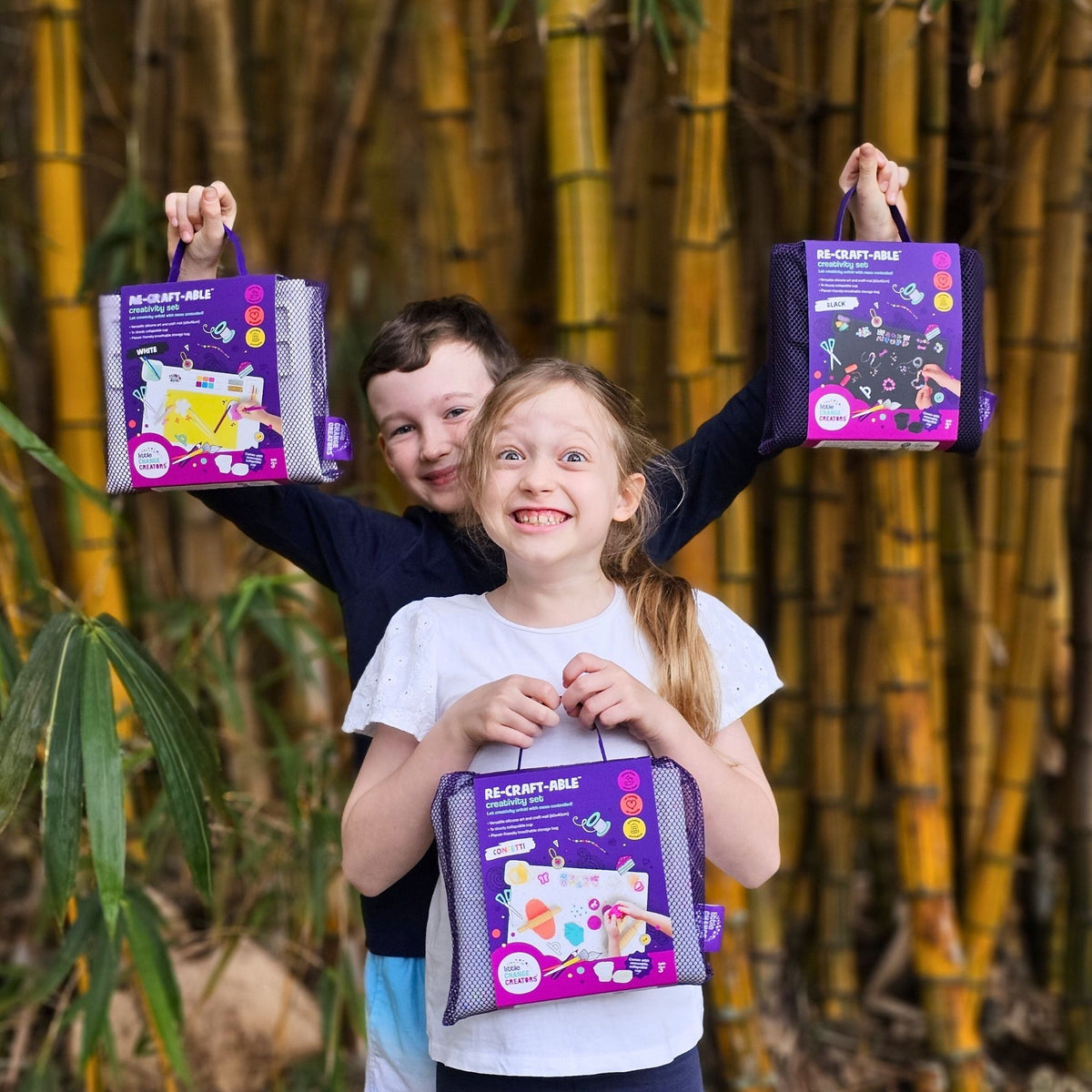 An excited boy and girl holding three Re-CRAFT-able Creativity Sets in purple packaging.