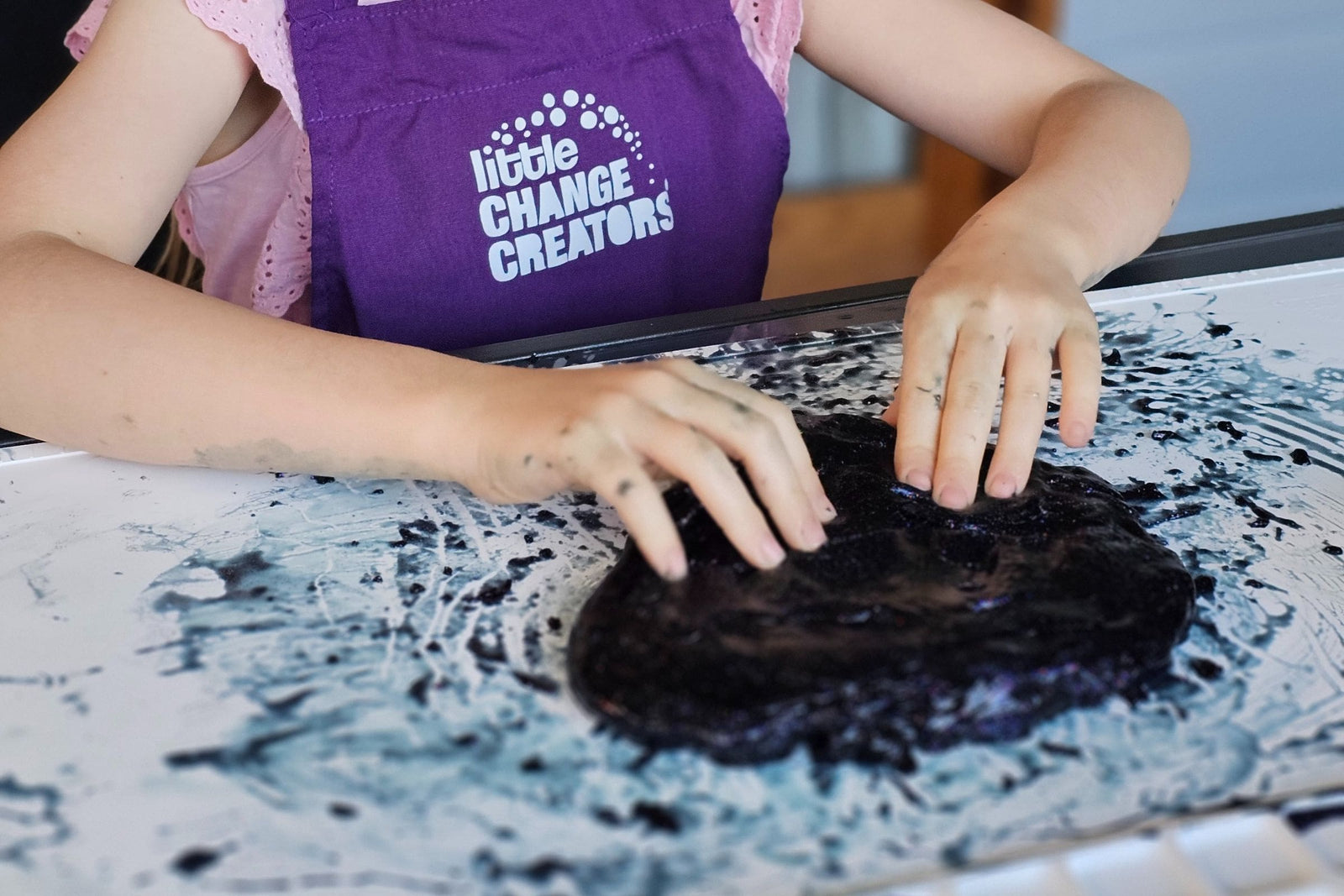 A child wearing a purple apron uses slime on a silicone craft mat.  The apron has a white Little Change Creators logo on its front panel.