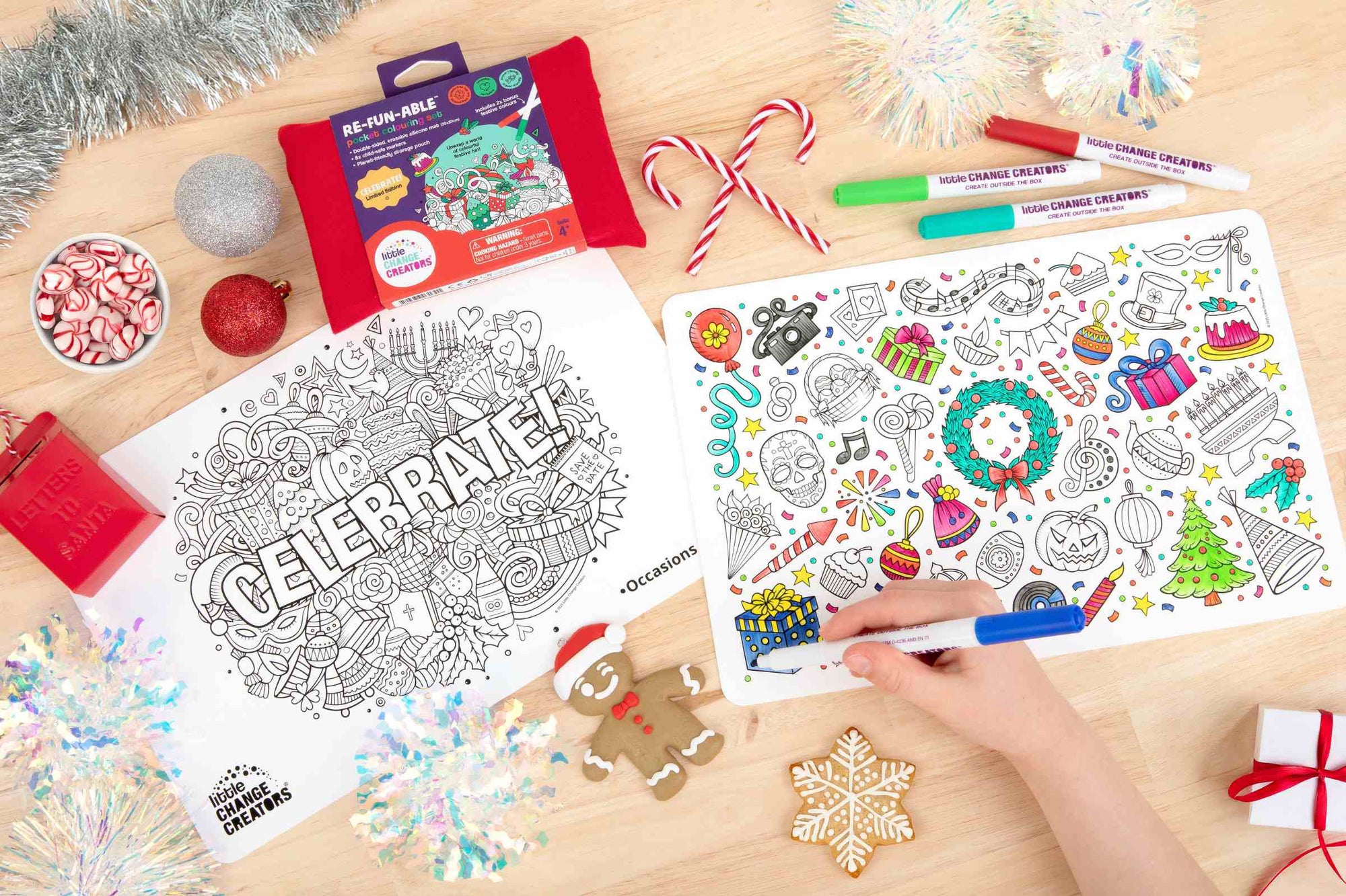 A wooden table top with two silicone colouring mats featuring festive illustrations.  The mats are surrounded by Christmas decorations including tinsel and candy canes.