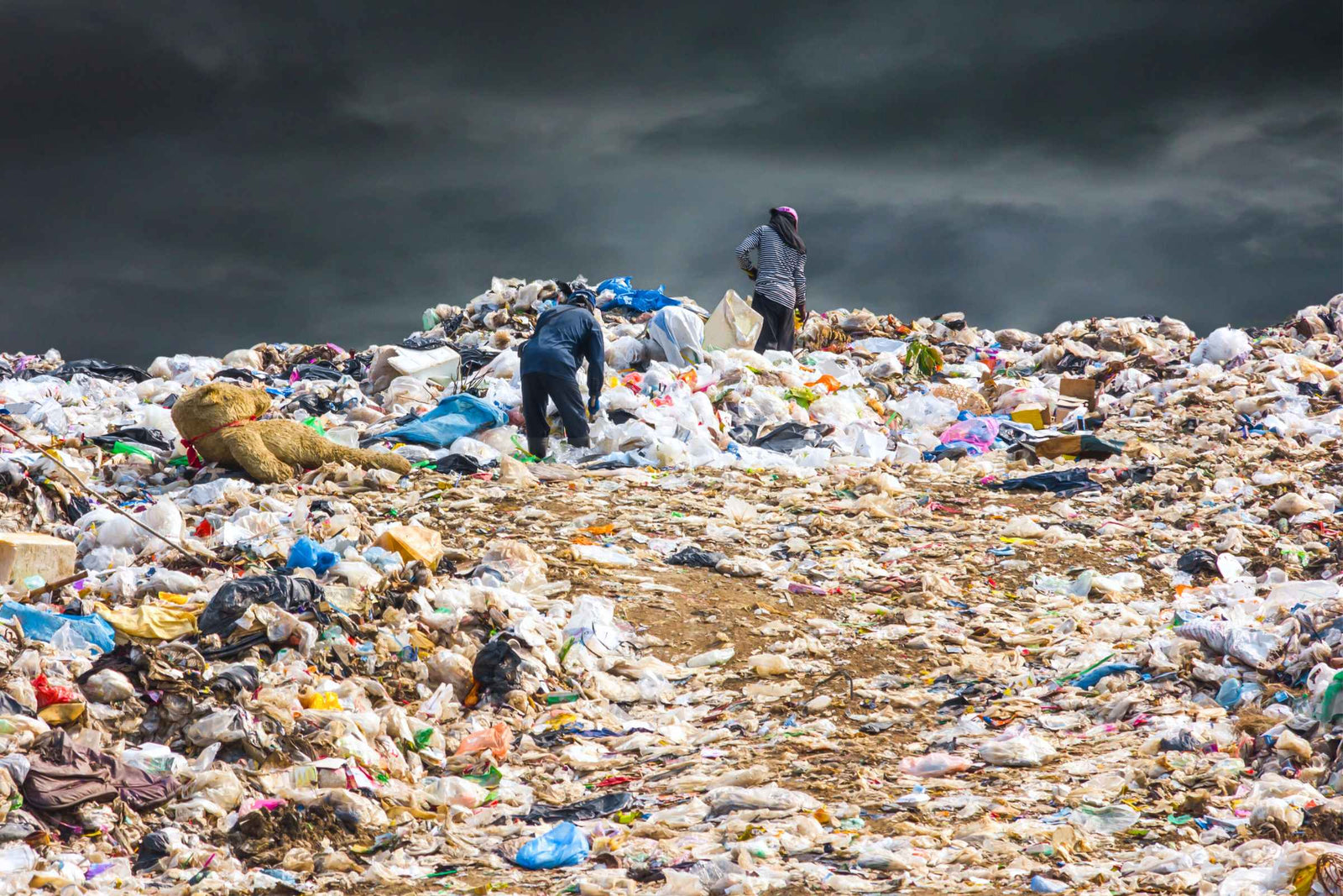 Landfill site showing packaging waste and the environmental impact of mass-consumerism.  Two people sift through the garbage against a dark, stormy sky.