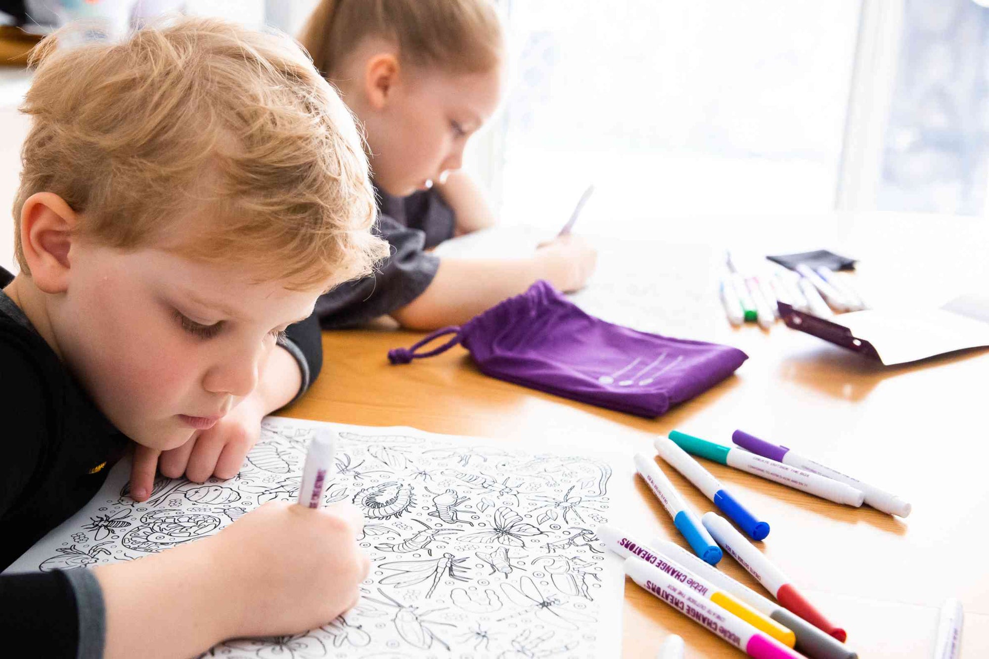 A well-lit mage of a boy and a girl sitting at a table drawing on a silicone colouring mat.  Colourful pens are sprawled across the table.