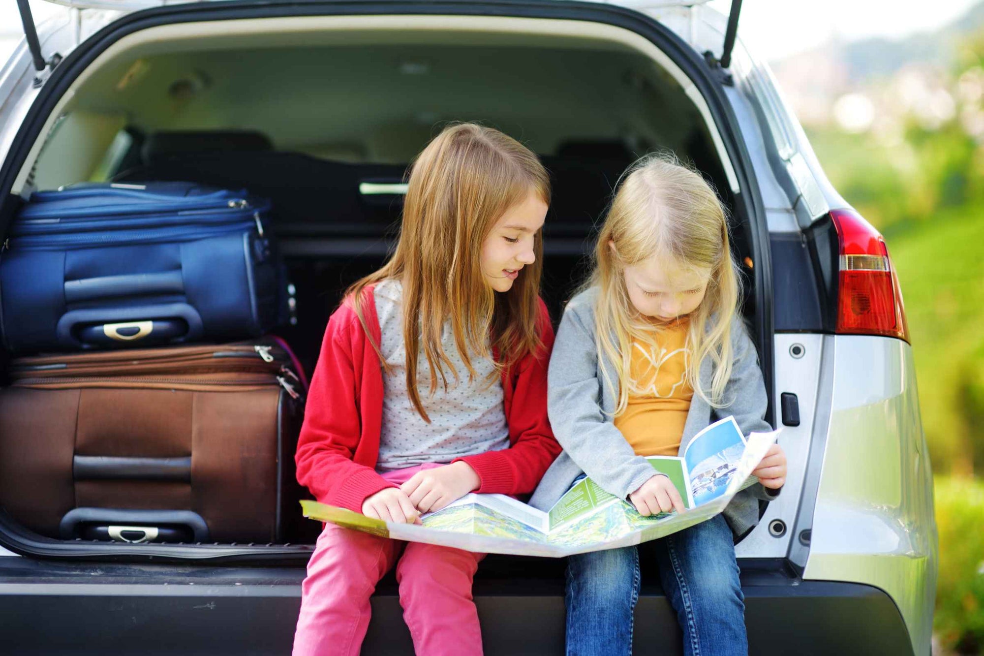 Image of two young girls sitting at the rear end of an open station wagon vehicle.  There are suitcases stacked inside the car and the girls are looking at a travel map.