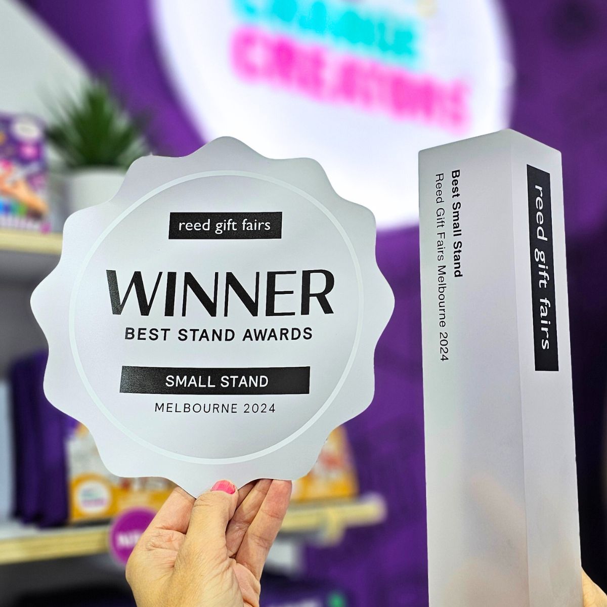 Image shows a silver rosette and trophy from Reed Gift Fairs for the Best Stand Awards at Melbourne 2024.  An illuminated sign with the Little Change Creators logo can be seen in the background.