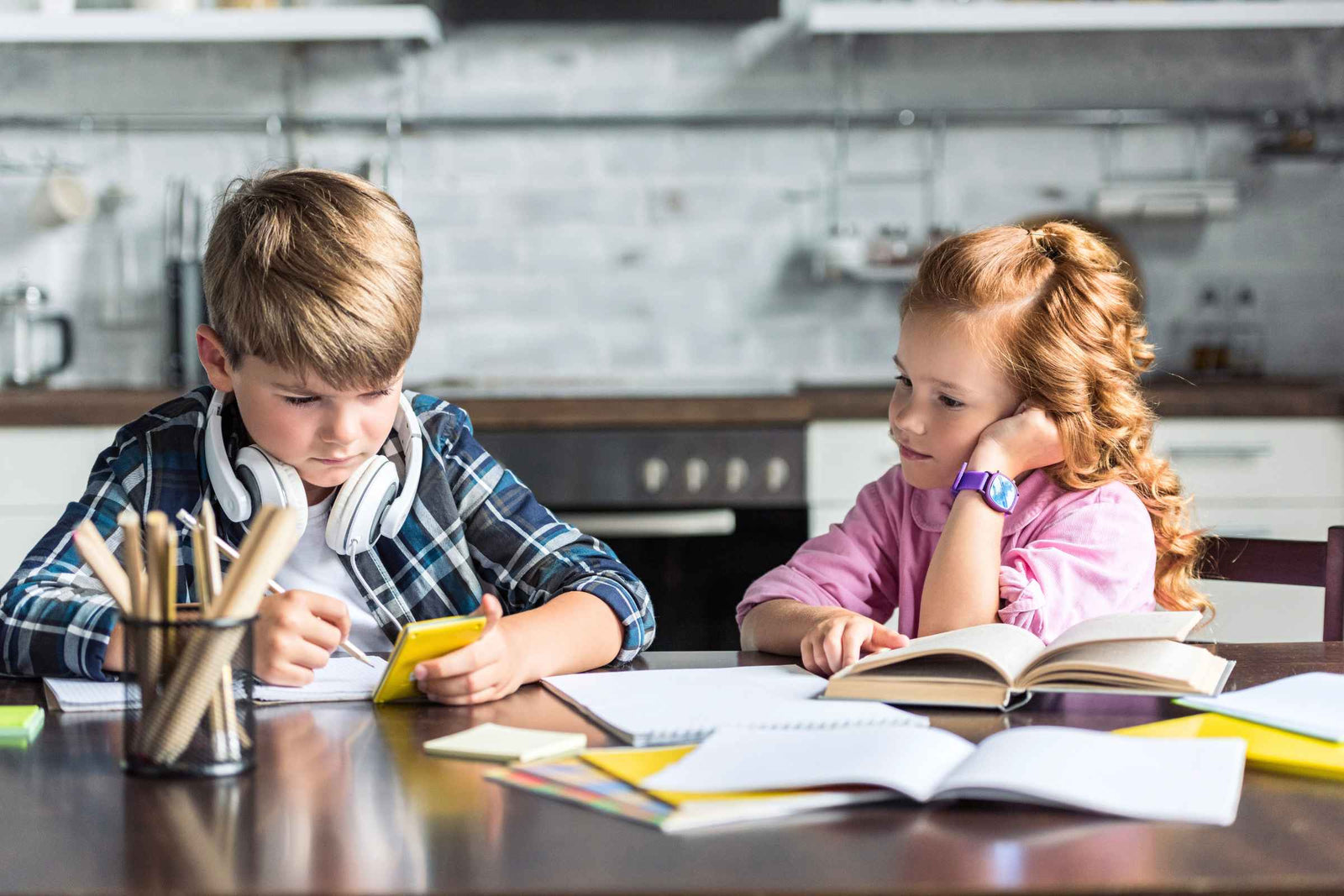 Two children engaged in screen-free activities, one reading a book and the other drawing, at a table with school supplies in a modern kitchen setting.