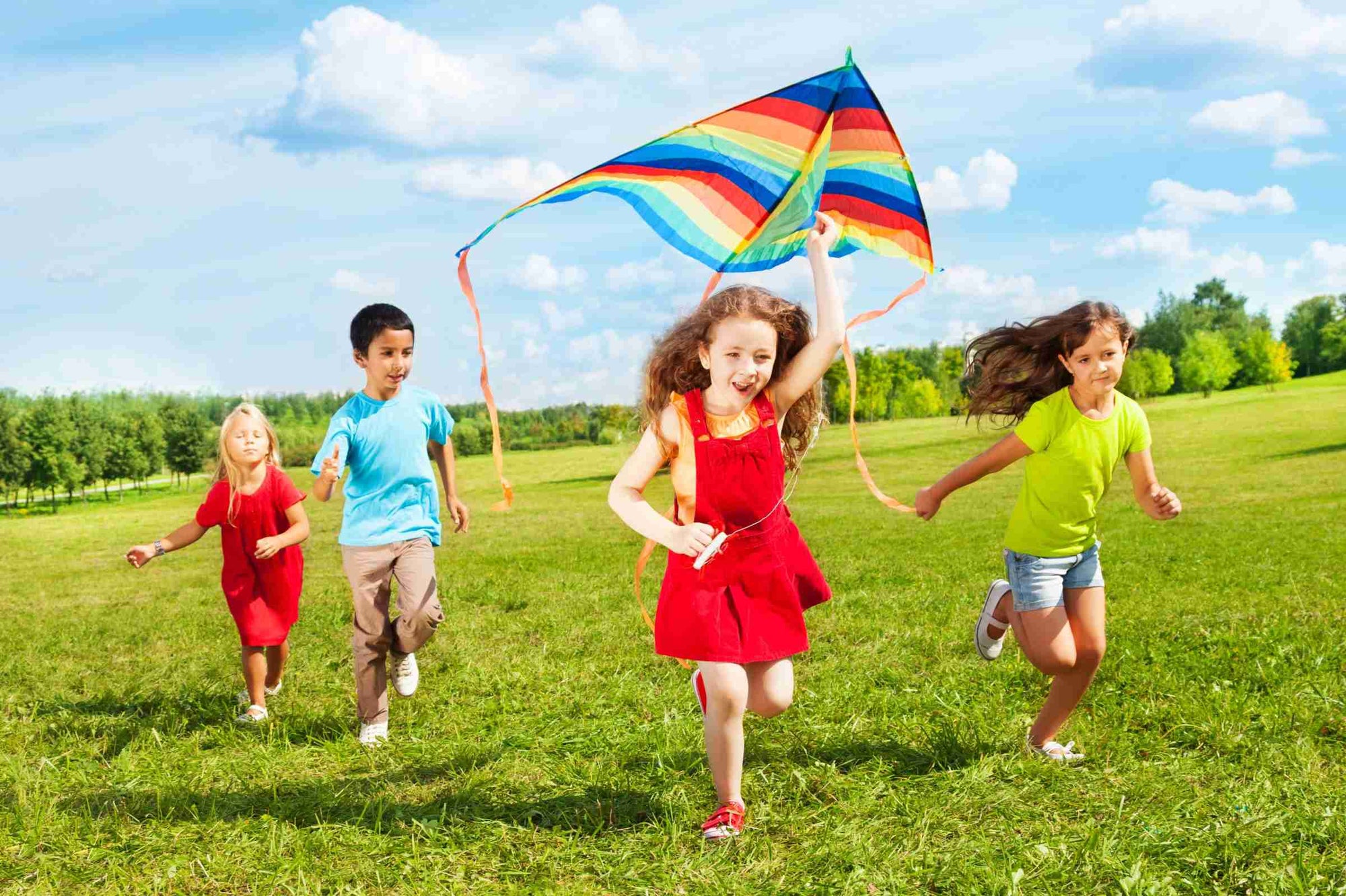 Colourful photo of children running across a green field with a rainbow kite.