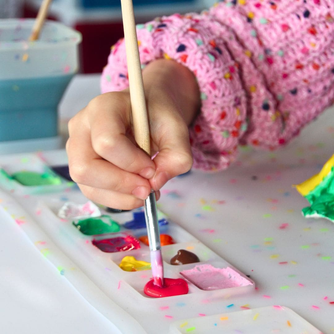 A child&#39;s hand using a paintbrush on a silicone painting mat with multi-coloured sprinkles.