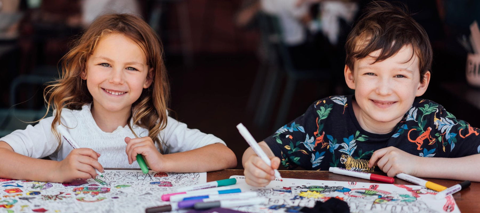 Two children smiling while colouring at a table with markers and silicone colouring mats in a real indoor setting.