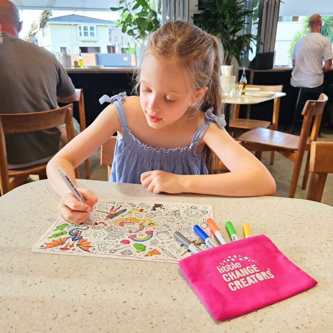 Image shows a girl using a Little Change Creators reusable silicone colouring mat at a cafe.  The mat features rainbows, unicorns, dragons and fairies.