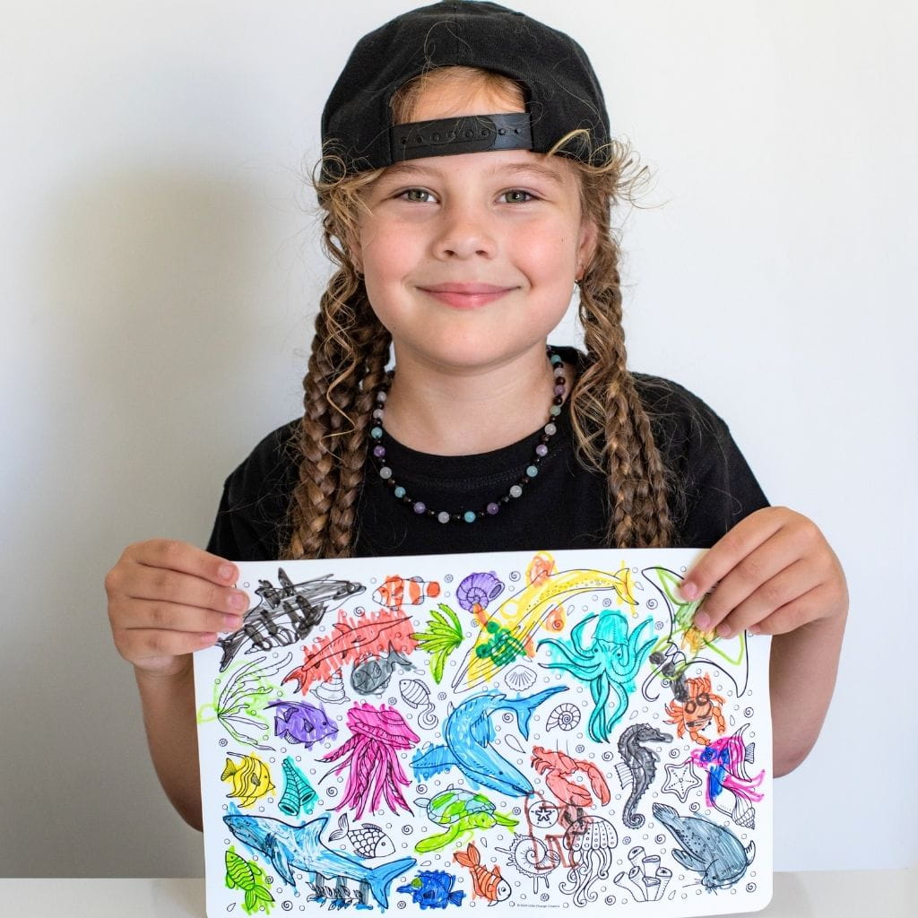 A young girl with braided hair, wearing a black cap and t-shirt, holds up a reusable silicone colouring mat featuring underwater sea life illustrations like dolphins, sharks, octopus, and jellyfish.