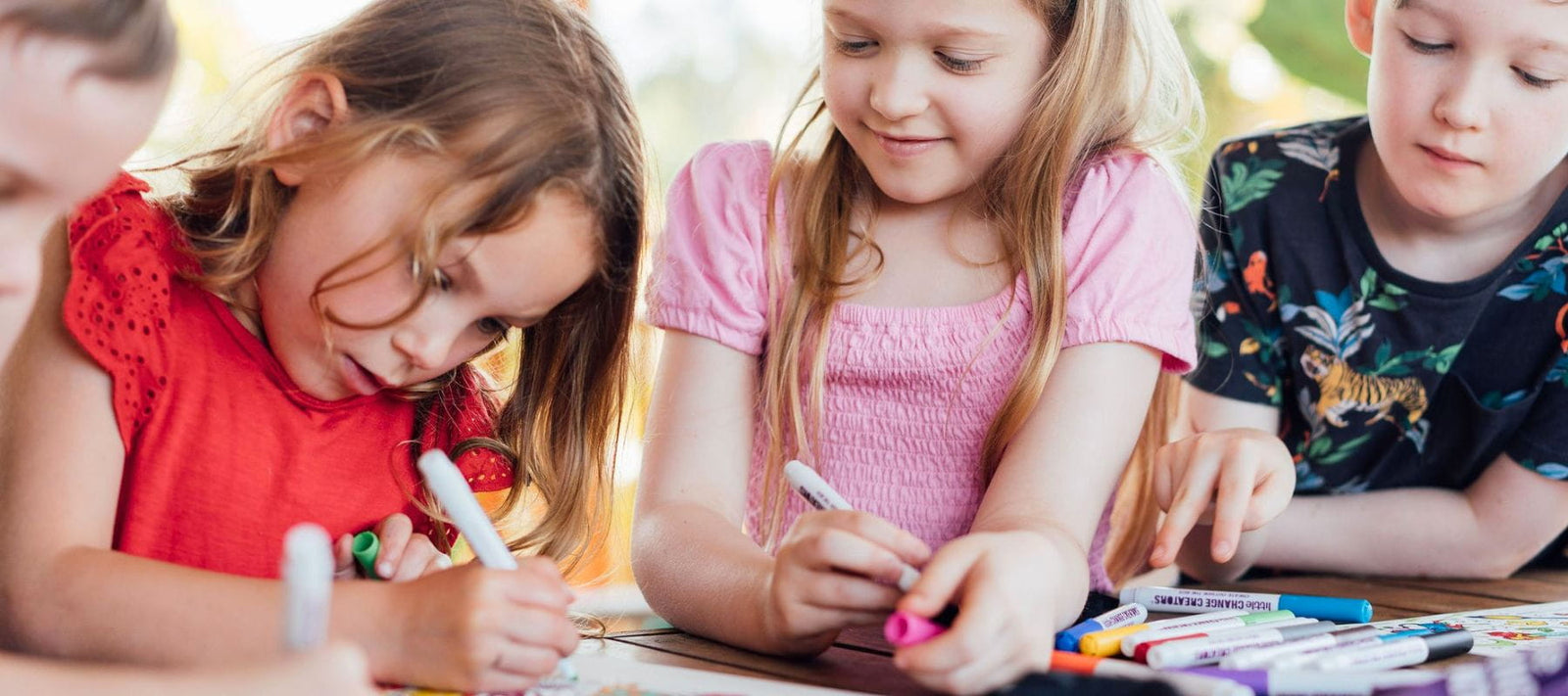 Children sitting at a table, drawing with markers on a silicone activity mat. The child in the center wears a pink top, the child on the left wears a red top, and the child on the right wears an animal print t-shirt.