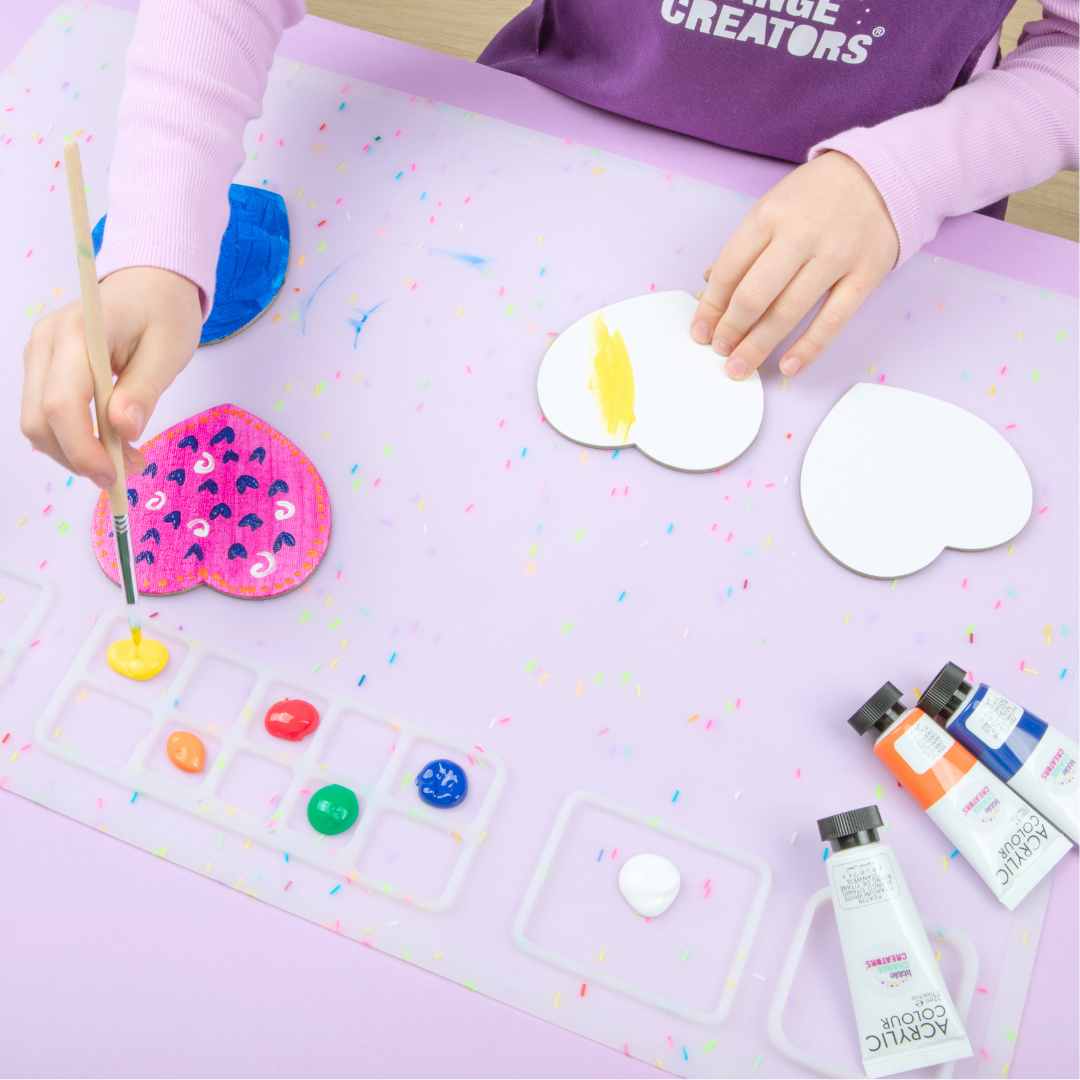 Child painting heart-shaped cards on a colourful silicone mat with art supplies around.