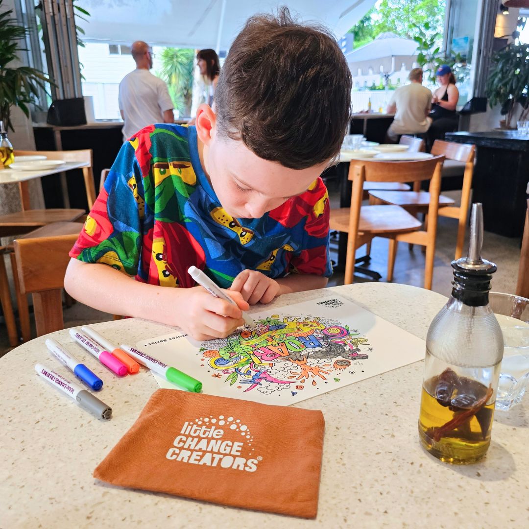 Image shows a boy colouring a reusable silicone doodle mat at a restaurant.  The drawing mat features dinosaurs and a volcano and comes with markers and a storage pouch.