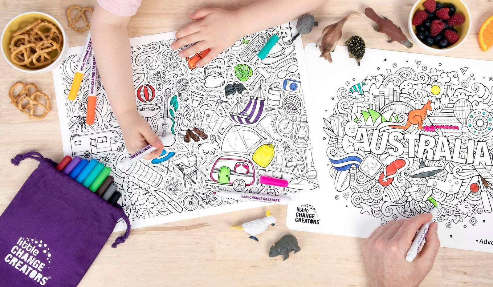 Adult and child seen from above colouring Outback and Australia reusable colouring sets with fruit, biscuits and animal toys on wooden table